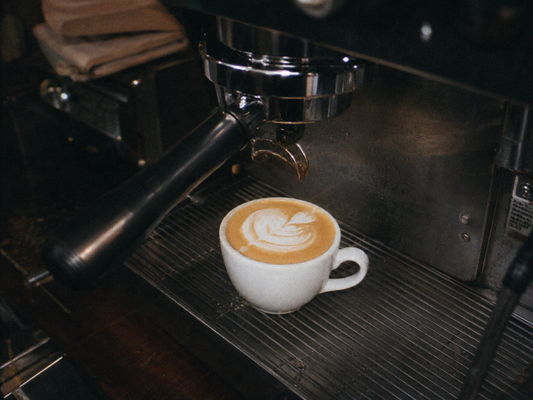 White latte cup with foam art sitting beneath an espresso machine in a local coffee shop.