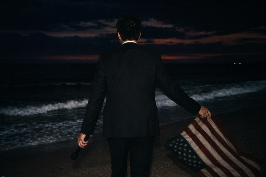 A cinematic scene of a man in a suit walking toward the ocean at dusk, holding a microphone in one hand and a loosely draped American flag in the other, symbolizing the intersection of politics, media, and advertising.