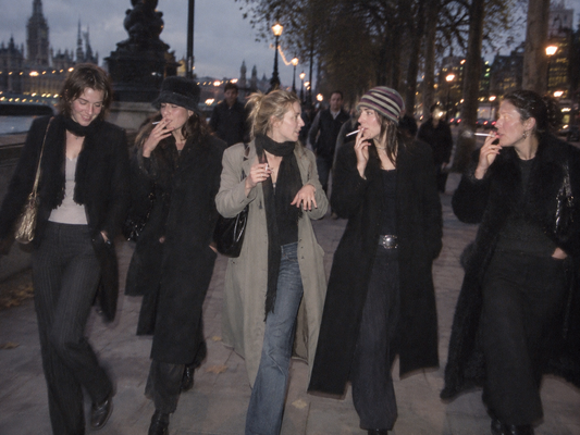 Five women walking together along a city sidewalk at dusk, wearing dark coats