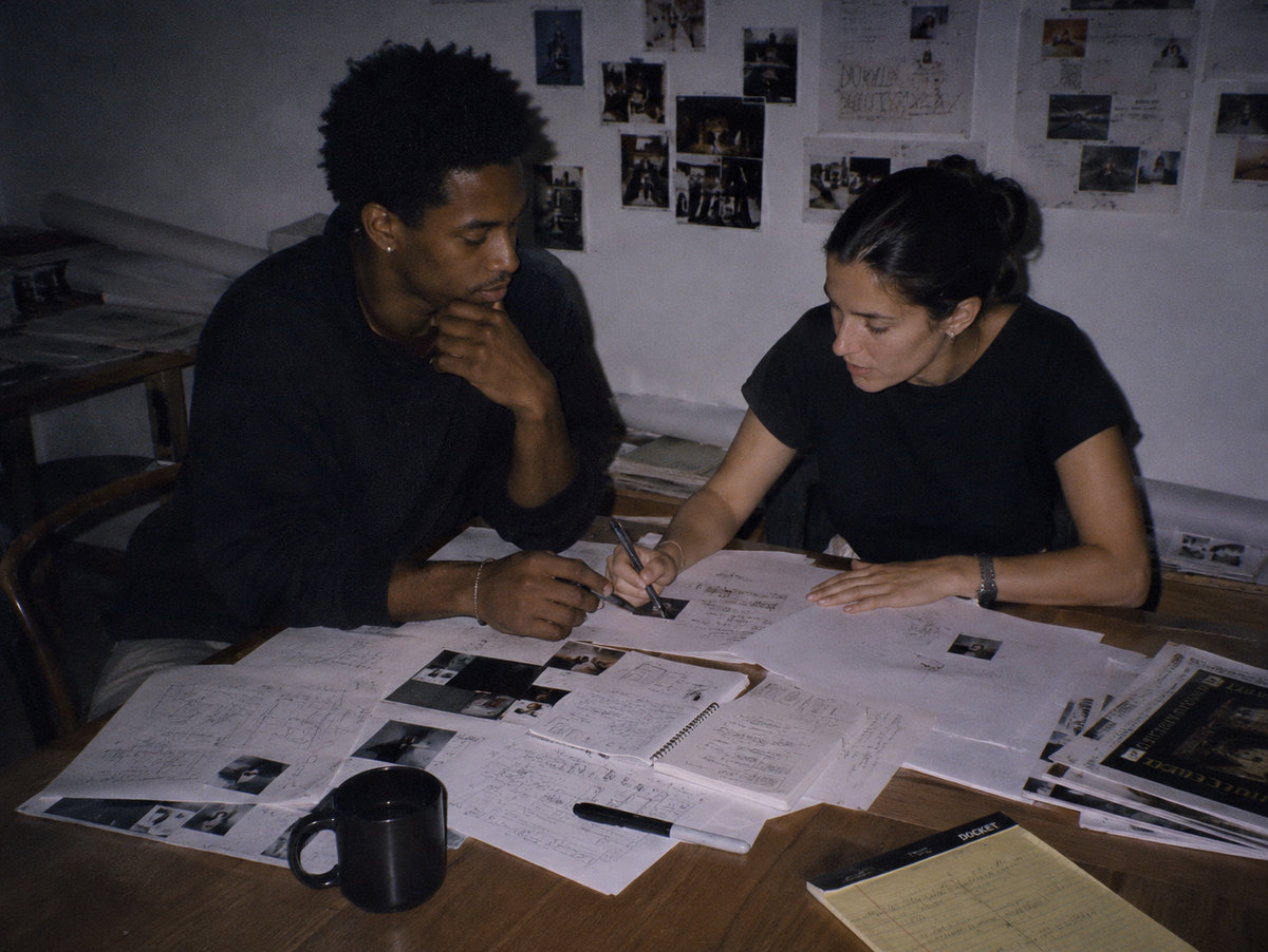 Two advertising creatives brainstorming ideas at a cluttered office table covered with sketches, notes, contact sheets, and campaign research.